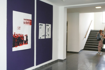 A large poster and two wall newspapers hang on a dark purple wall in a university hallway.
