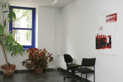 A large poster hangs in a secluded hallway of the university. Next to it are two office chairs and two large plants, as if forgotten.