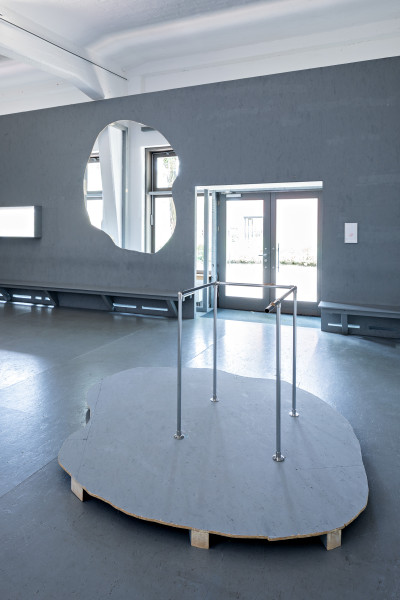 Eine Installation aus Metallstangen, die einen Stand bilden, steht auf einer freiförmigen grauen Holzebene. Über sie hinweg schaut man auf eine raumhohe graue Wand, aus der ebendiese Freiform herausgeschnitten ist. Durch das Loch sowie die Eingangstür fällt helles Tageslicht in den Raum.
