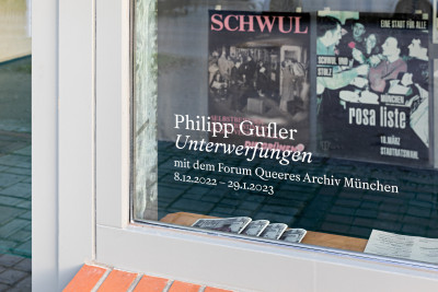 Auf einer Fensterscheibe steht der Titel der Ausstellung, mit einem Verweis auf die Laufzeit. Hinter dem Fenster liegen Hefte und hängen zwei Plakate.