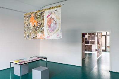 In a high room, a colorful quilt hangs from the ceiling. Below it is a table with two gray stools. On it lie various notebooks and books. Through a doorway you can look into the neighboring room and see poster walls.