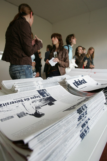 Folded newspapers lie in high piles on a table. Young people are standing behind them, talking.