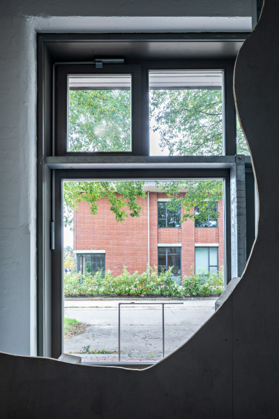 A view through the recess of a gray wall to the outside. In front of the window is a sculpture made of narrow metal bars. Behind it is a street and a brick building.