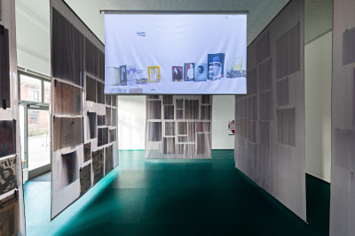 Auf einer hängenden Leinwand spielt ein Video. Um sie herum hängen graue Stoffbahnen, die einen Raum erschaffen. Schattenwürfe lassen Poster erahnen, die sich auf der Außenseite der Wände befinden.