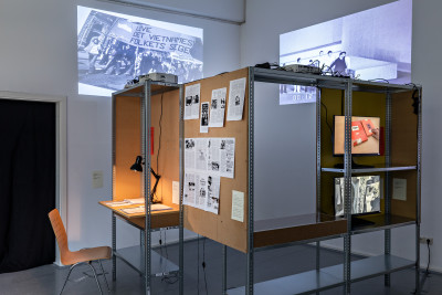 In a shelf system, two screens show video works, texts are displayed on a reading table and others hang on a pinboard. Two black-and-white videos play on the wall behind them.