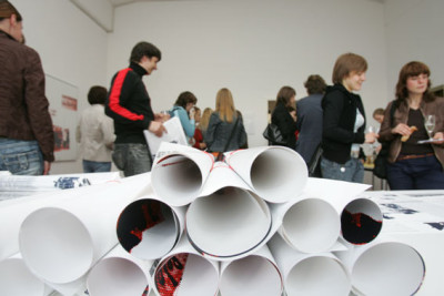Many young people are standing in small groups, talking and drinking champagne. In the foreground, rolls of posters are piled up on a table.